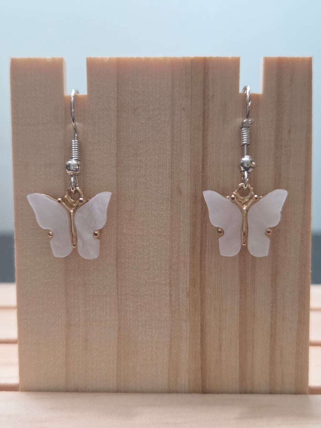 Close-up of mother-of-pearl shell butterfly earrings with gold-tone bail and silver fish hook ear wires, showing natural iridescent shimmer on white wings