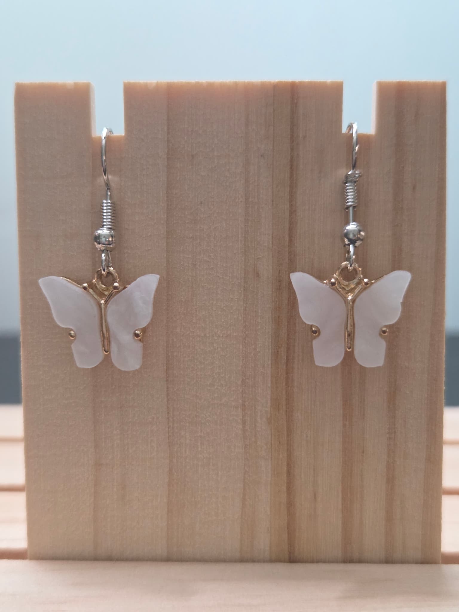 Close-up of mother-of-pearl shell butterfly earrings with gold-tone bail and silver fish hook ear wires, showing natural iridescent shimmer on white wings