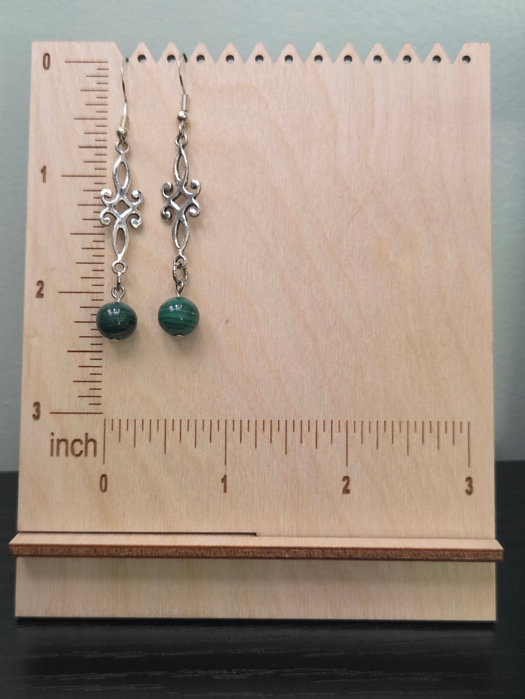 Malachite scroll drop earrings displayed on a wooden earring stand with ruler showing approximately 2 1/8 inch length