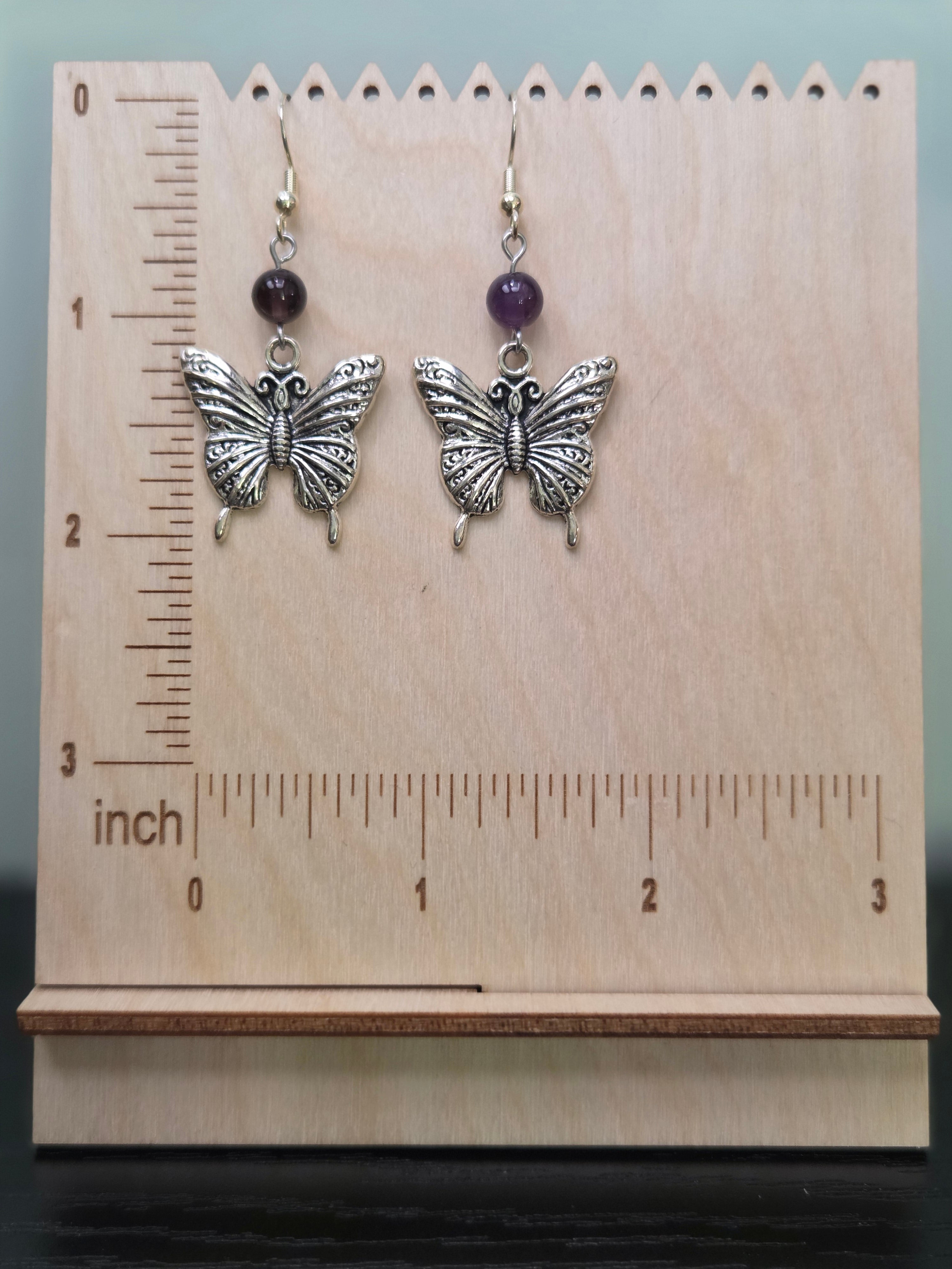 Butterfly amethyst drop earrings displayed on a wooden earring stand with ruler showing approximately 2 inch length