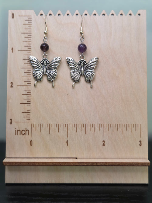 Butterfly amethyst drop earrings displayed on a wooden earring stand with ruler showing approximately 2 inch length