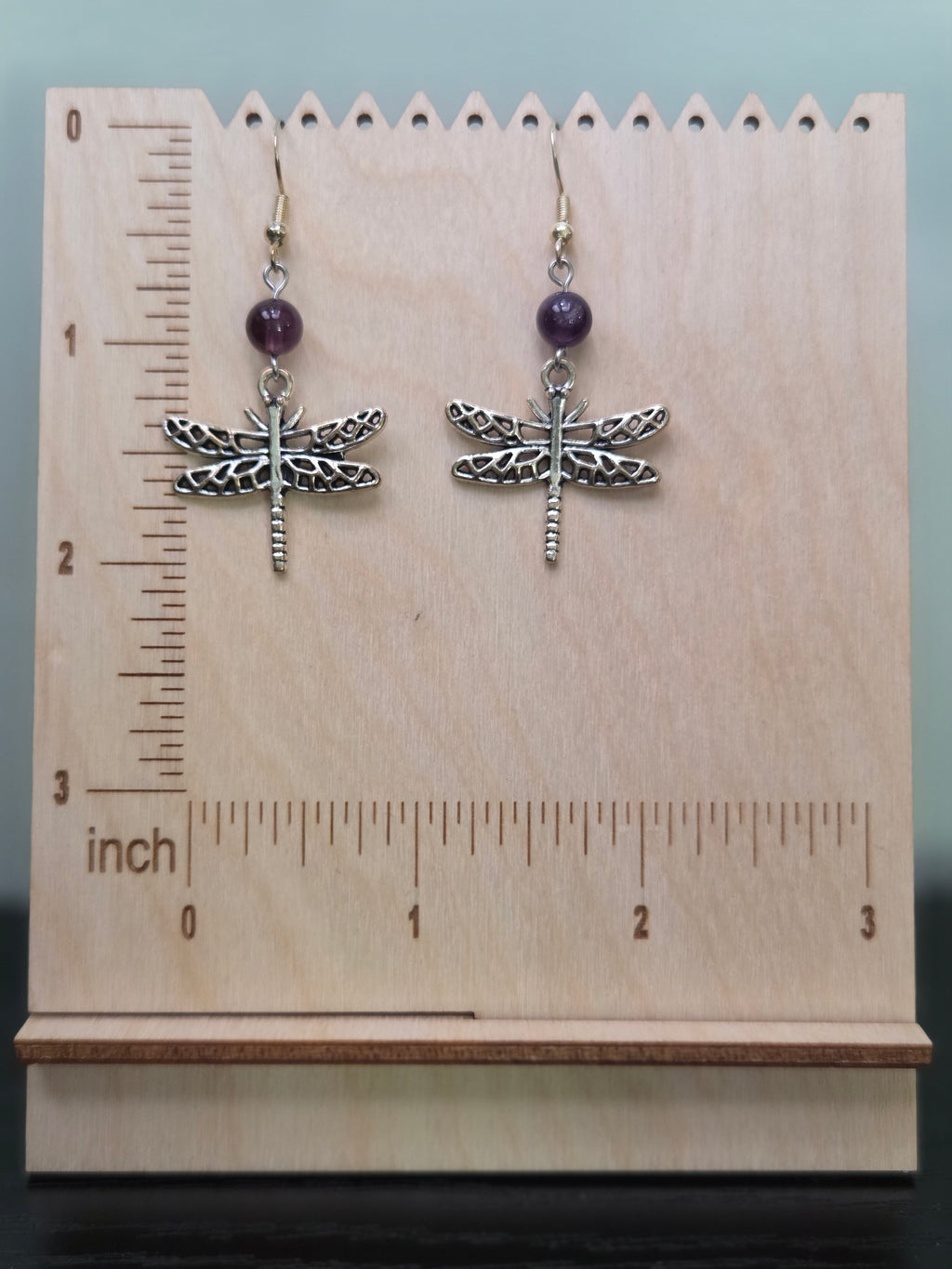 Dragonfly amethyst drop earrings displayed on a wooden earring stand with ruler showing approximately 2 inch length