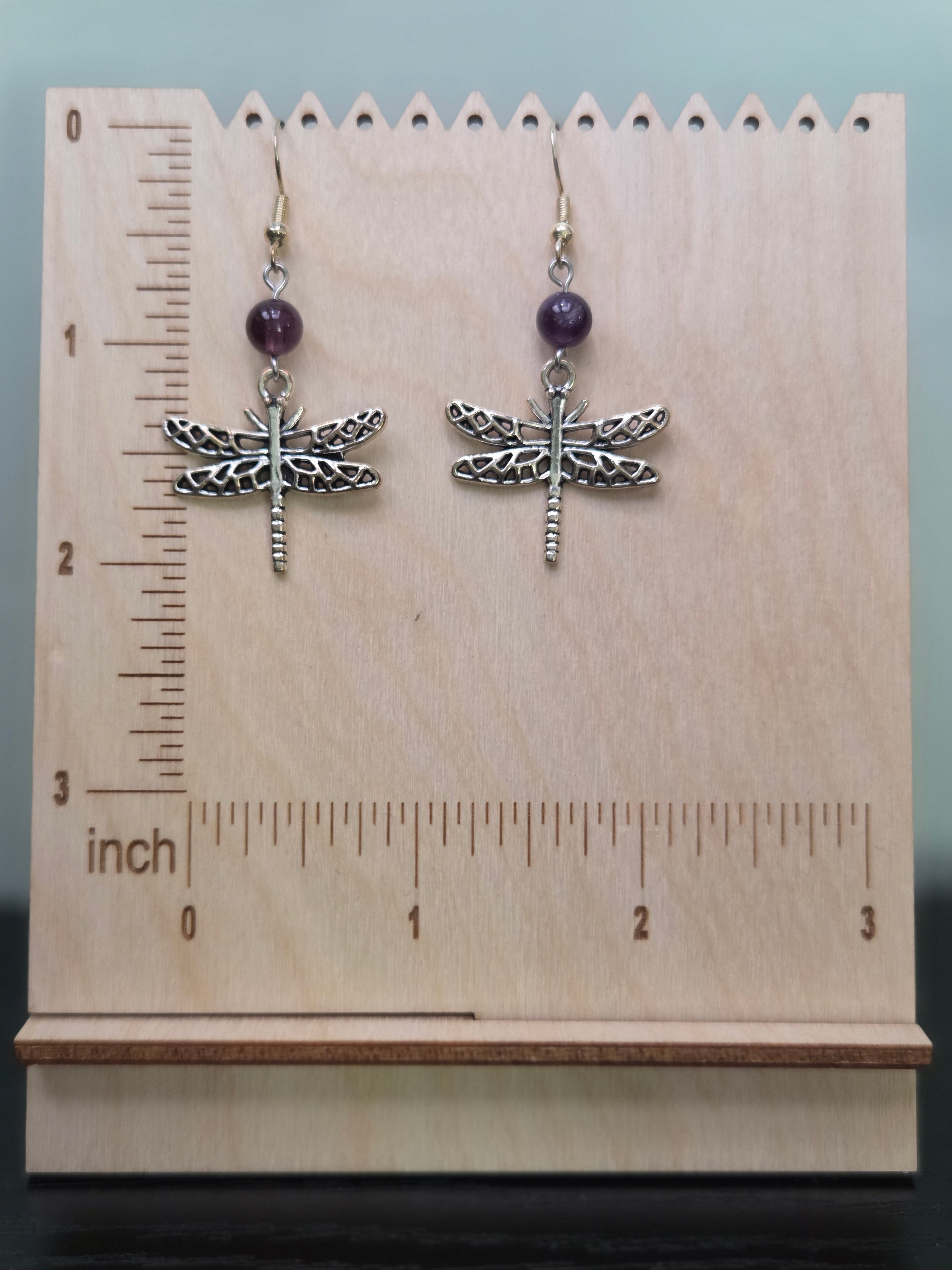 Dragonfly amethyst drop earrings displayed on a wooden earring stand with ruler showing approximately 2 inch length