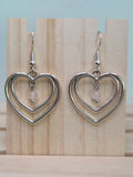 Silver-tone double heart earrings with a rose quartz bead dangling at center, close-up on wood block