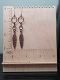 Cascading feather earrings with two reclaimed copper-tone connector rings above a feather charm, shown on a wooden ruler stand measuring approximately 2.75 inches