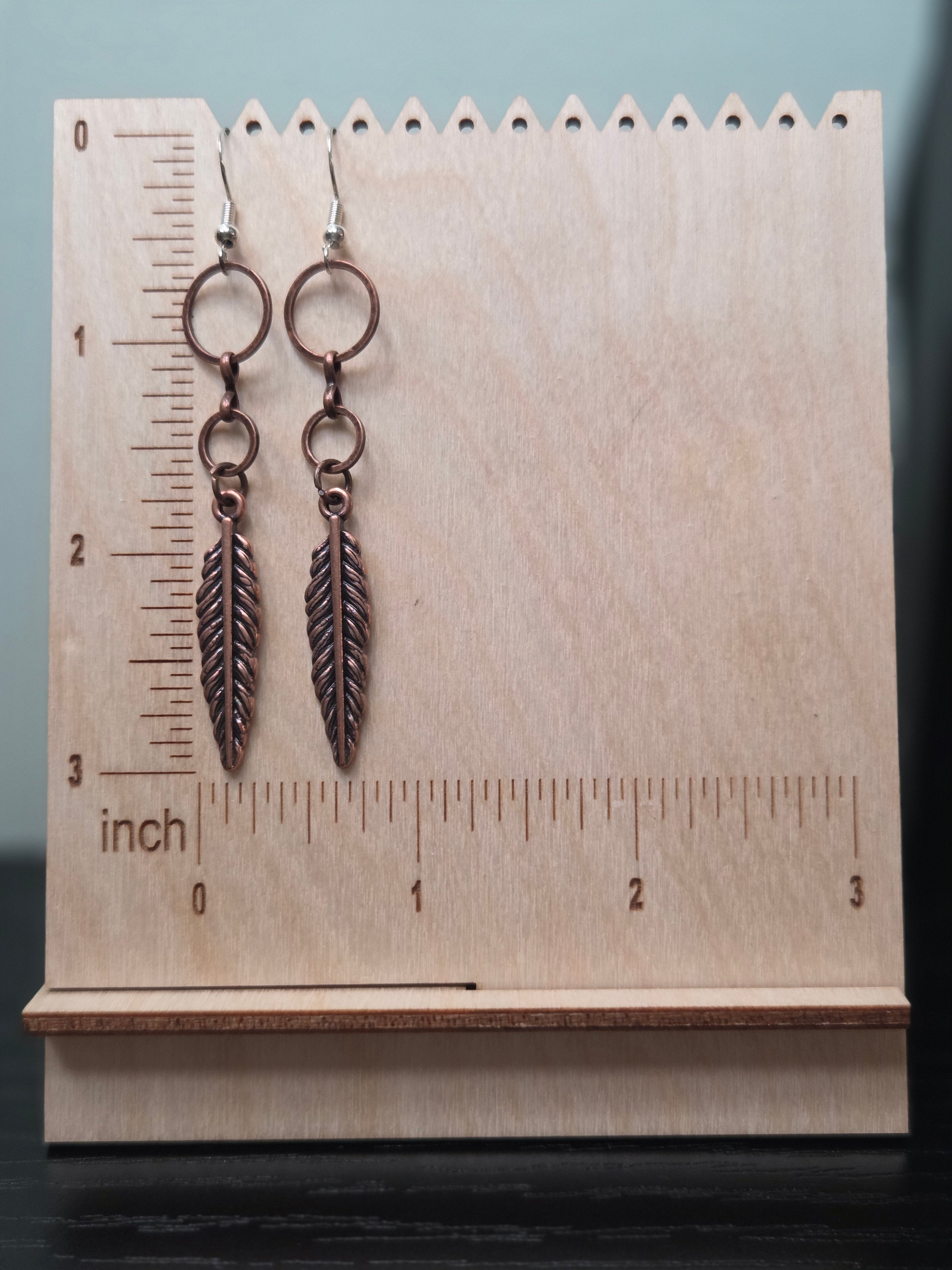 Cascading feather earrings with two reclaimed copper-tone connector rings above a feather charm, shown on a wooden ruler stand measuring approximately 2.75 inches