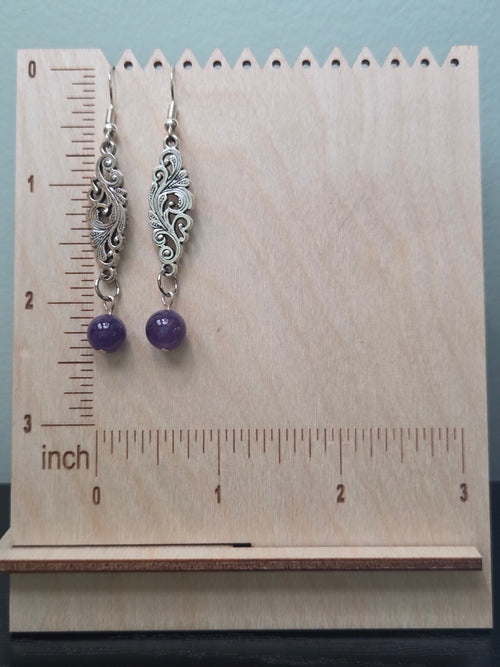 Close-up of handmade filigree amethyst drop earrings showing antique silver scrollwork connector and round purple amethyst bead on pink background