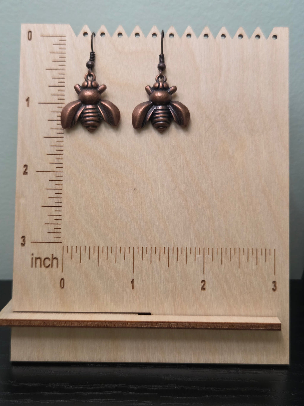 Close-up of bronze bee charm earrings in natural lighting on kraft card display, showing detailed wing texture, segmented body, and warm copper-brown metallic tone