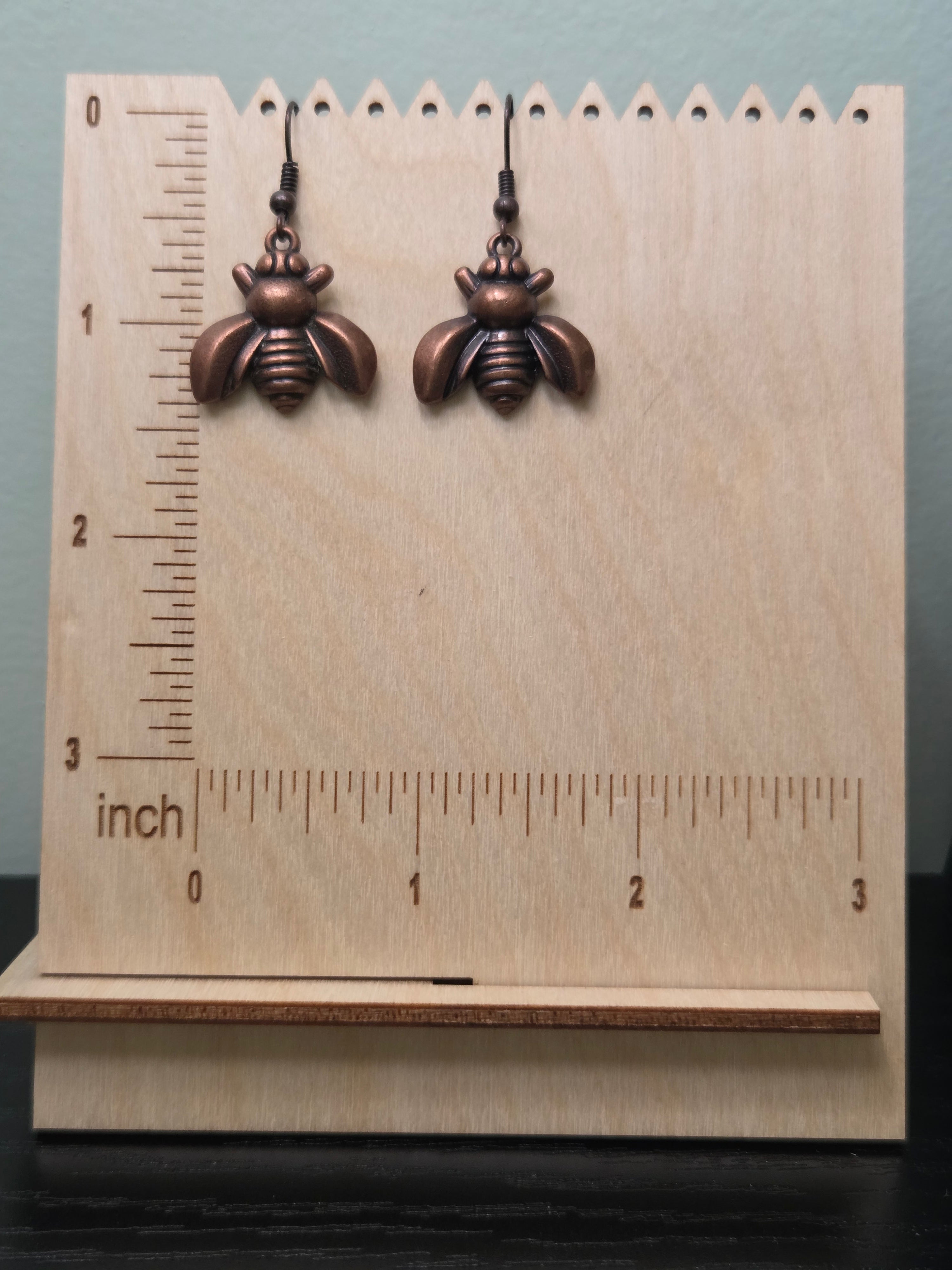 Close-up of bronze bee charm earrings in natural lighting on kraft card display, showing detailed wing texture, segmented body, and warm copper-brown metallic tone