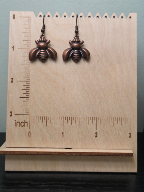 Close-up of bronze bee charm earrings in natural lighting on kraft card display, showing detailed wing texture, segmented body, and warm copper-brown metallic tone