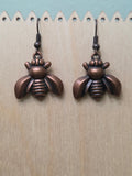 Bronze bee charm dangle earrings displayed on a wooden ruler stand, showing full length of approximately 1 inch, warm copper-tone metallic finish with detailed wings and body