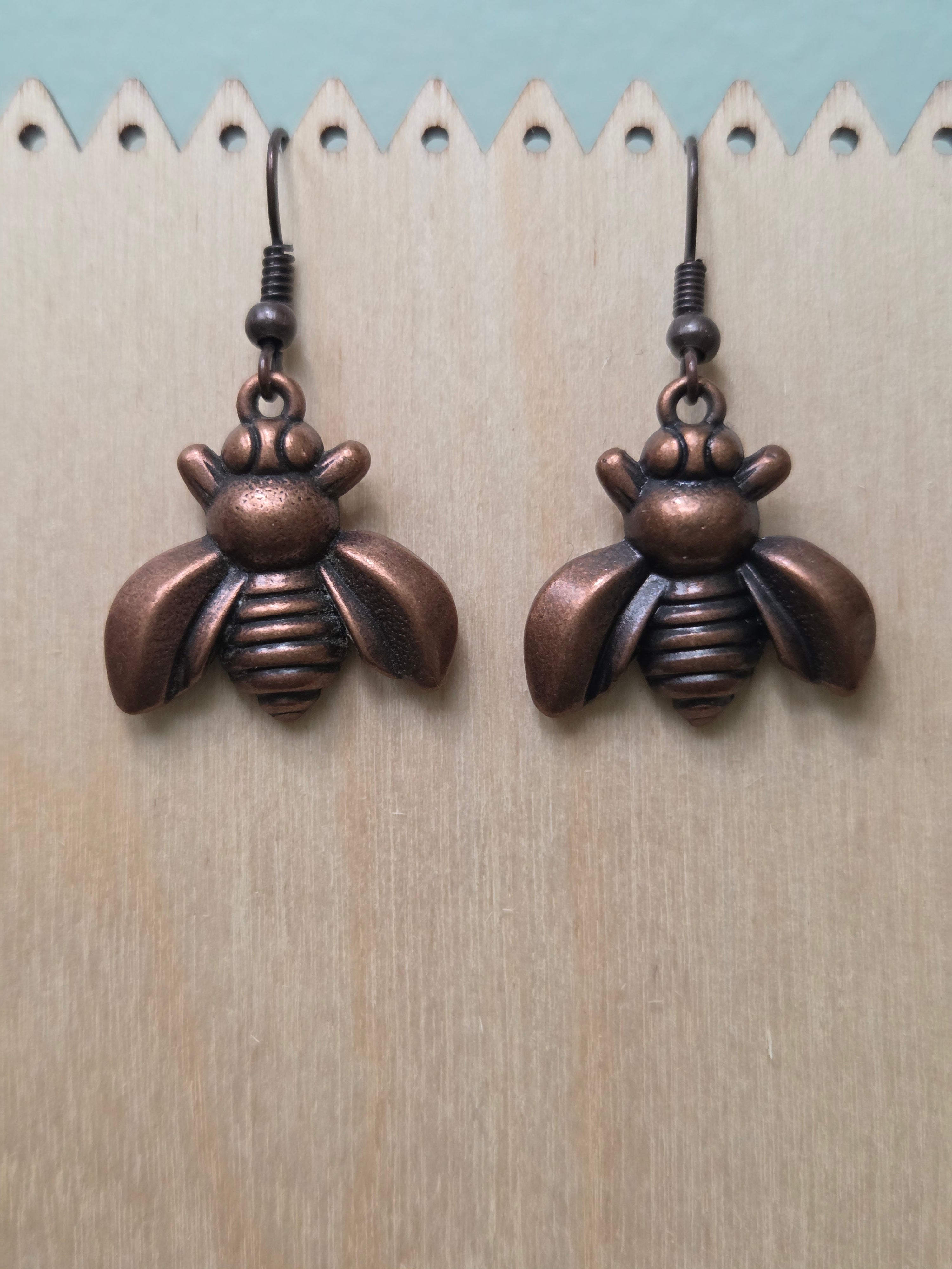 Bronze bee charm dangle earrings displayed on a wooden ruler stand, showing full length of approximately 1 inch, warm copper-tone metallic finish with detailed wings and body