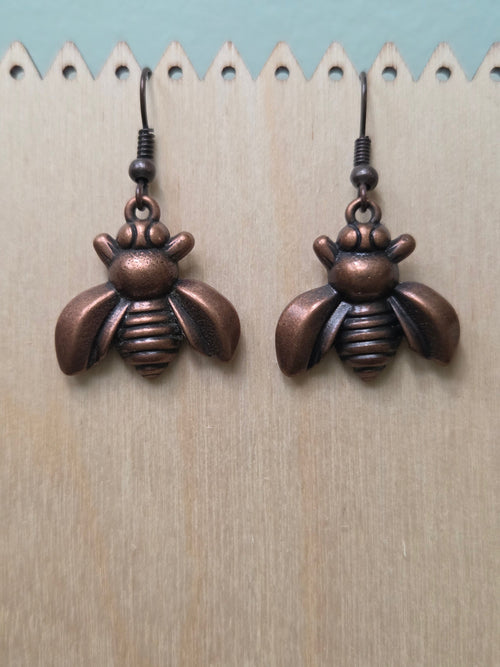 Bronze bee charm dangle earrings displayed on a wooden ruler stand, showing full length of approximately 1 inch, warm copper-tone metallic finish with detailed wings and body
