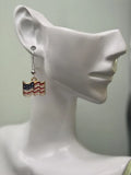 Handmade waving American flag earring on a white mannequin ear, featuring bold red and white wavy stripes with a navy blue star field and cream border in a gold-tone frame