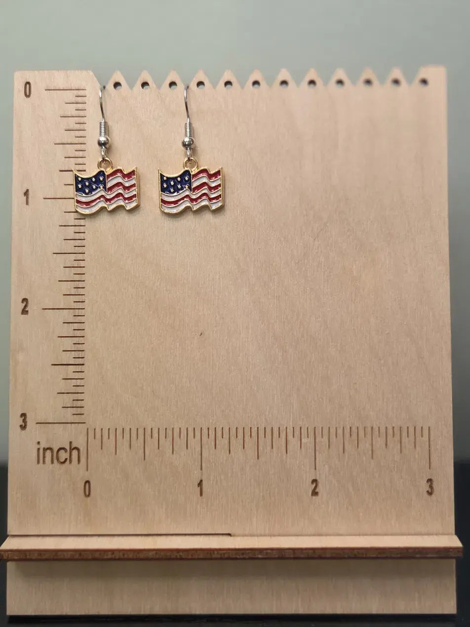 Pair of waving American flag dangle earrings on a wooden display board with ruler showing approximately 1 inch charm size, featuring red and white wavy stripes with a navy blue star field and cream-toned border in a gold-tone frame