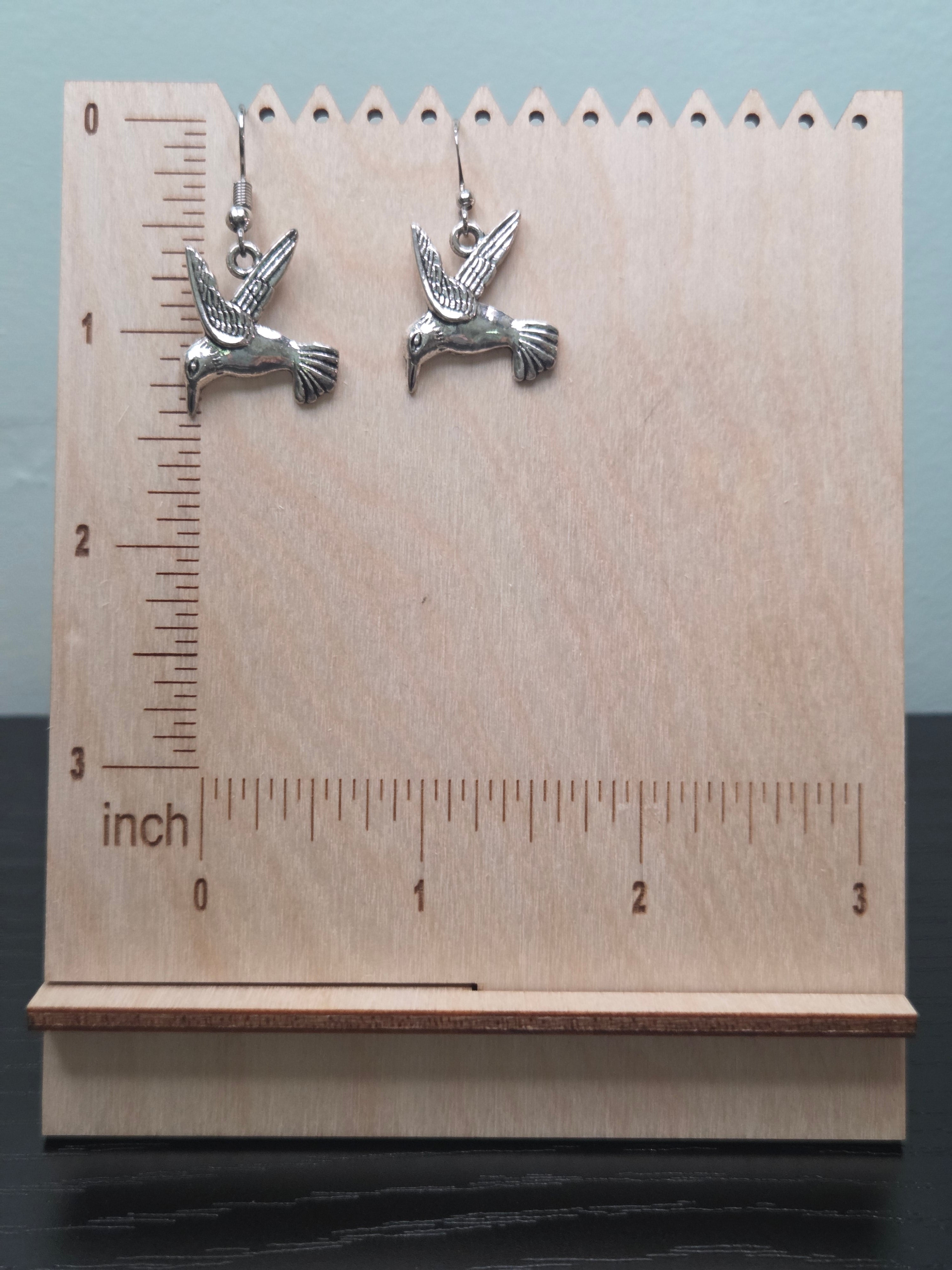 Silver-tone alloy hummingbird dangle earrings against ruler showing approximately 1 inch charm length