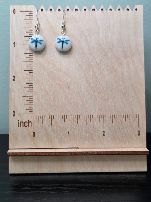 Pair of white round glass bead earrings with blue dragonfly design on wooden ruler display, showing approximately 1 inch drop length, silver lever-back hooks, natural lighting

