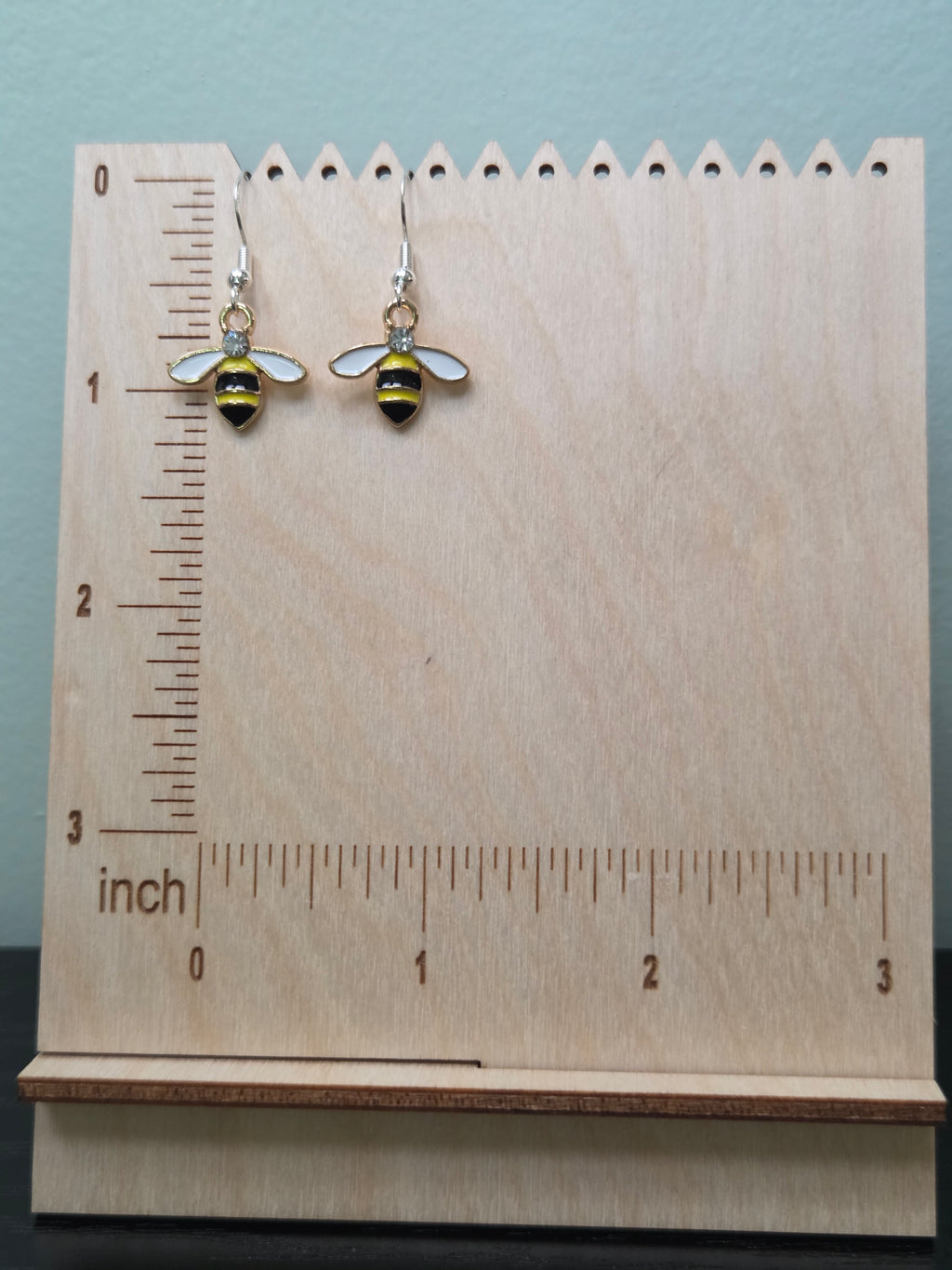  Pair of bumble bee dangle earrings with yellow and black enamel body, white enamel wings, rhinestone accent, and silver-tone fishhook wires, displayed on a wooden earring stand with inch ruler for scale — charm measures approximately 1 inch