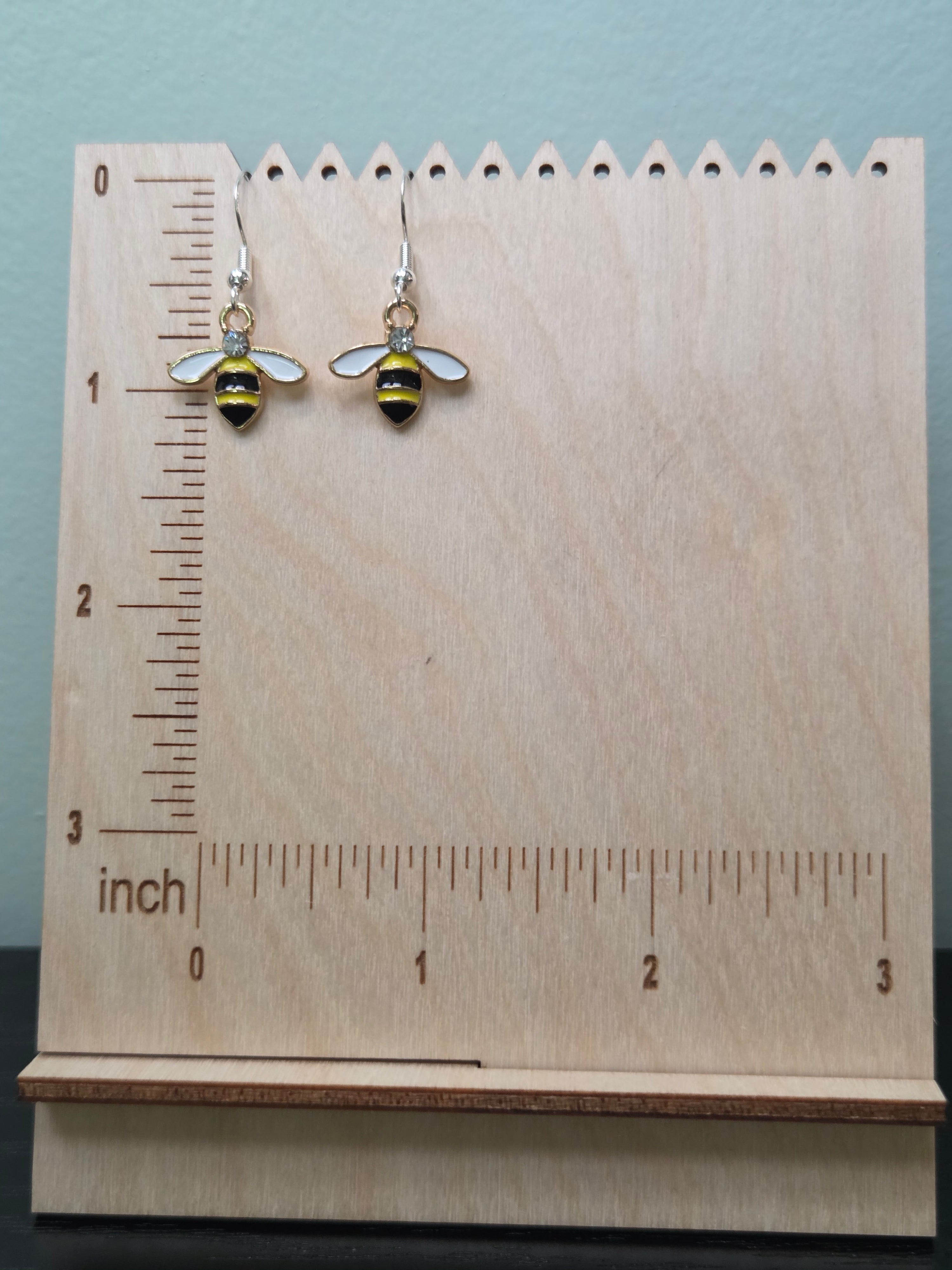  Pair of bumble bee dangle earrings with yellow and black enamel body, white enamel wings, rhinestone accent, and silver-tone fishhook wires, displayed on a wooden earring stand with inch ruler for scale — charm measures approximately 1 inch