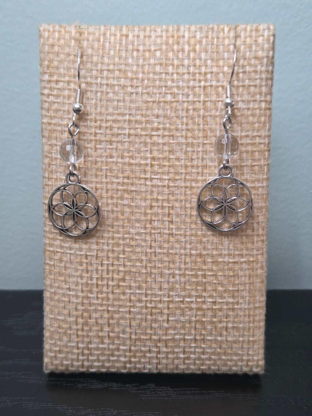 Small Seed of Life sacred geometry charm earrings with clear quartz bead, silver-tone, shown on wood display