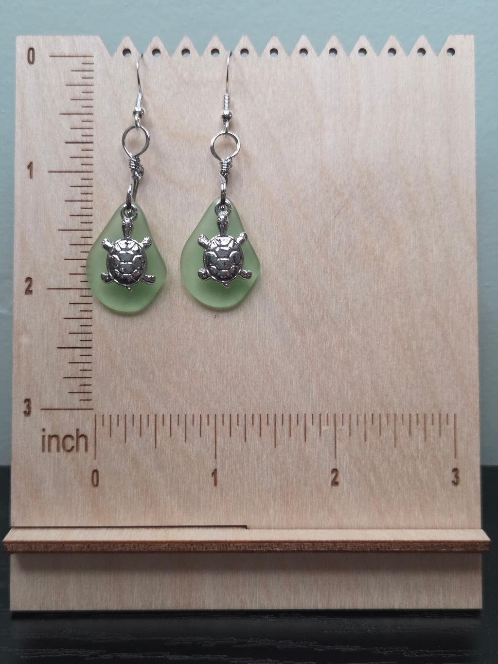 Pair of sage green frosted glass turtle charm earrings displayed on a wooden ruler stand showing approximately 1.75 inch length