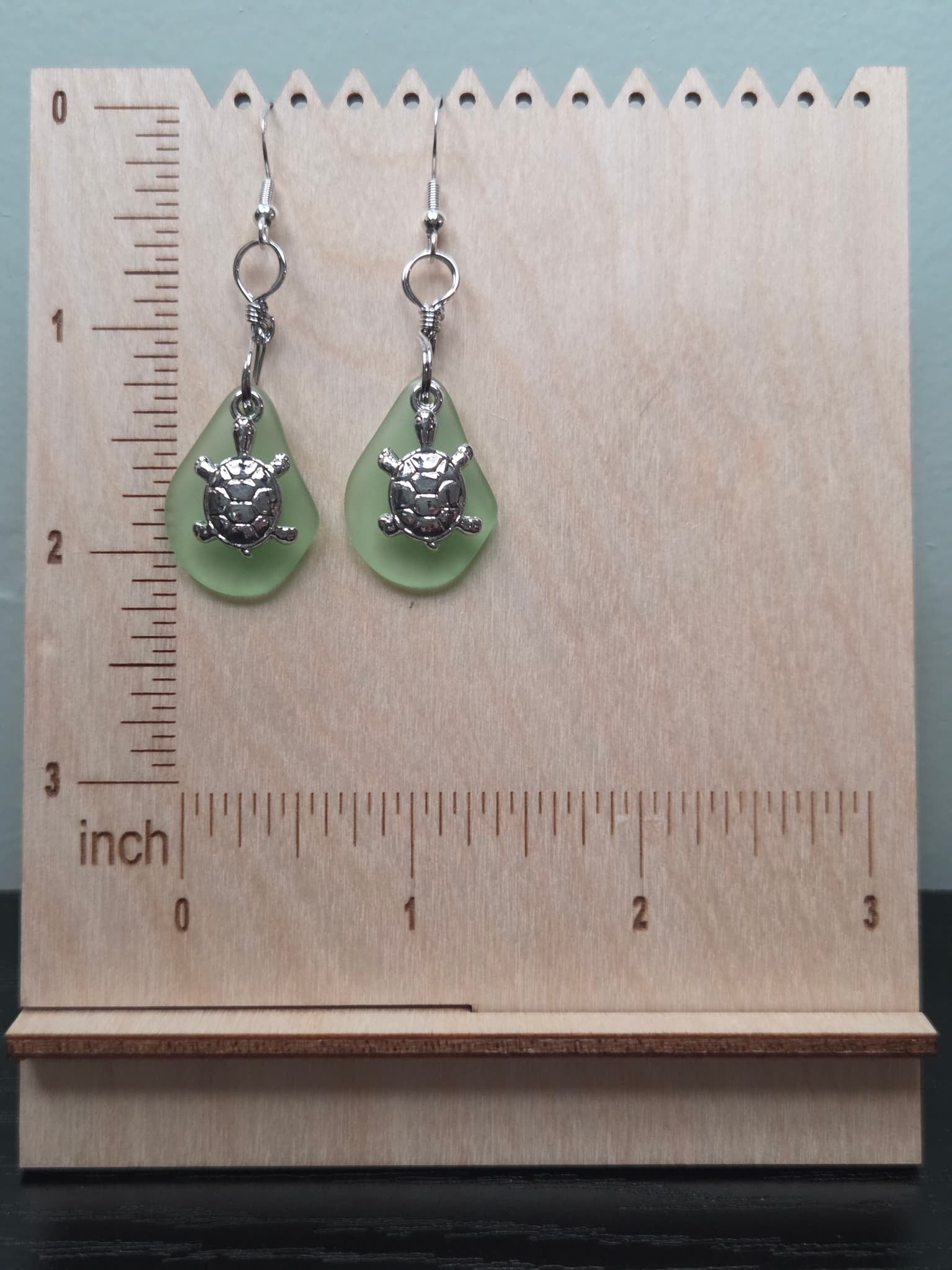 Pair of sage green frosted glass turtle charm earrings displayed on a wooden ruler stand showing approximately 1.75 inch length