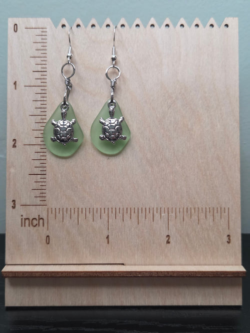 Pair of sage green frosted glass turtle charm earrings displayed on a wooden ruler stand showing approximately 1.75 inch length