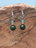 Pair of dangle earrings featuring a deep green jade round bead below a lighter sage green aventurine bead, joined by a gold-tone accent bead, on silver-tone fish hook ear wires, resting on a piece of natural wood