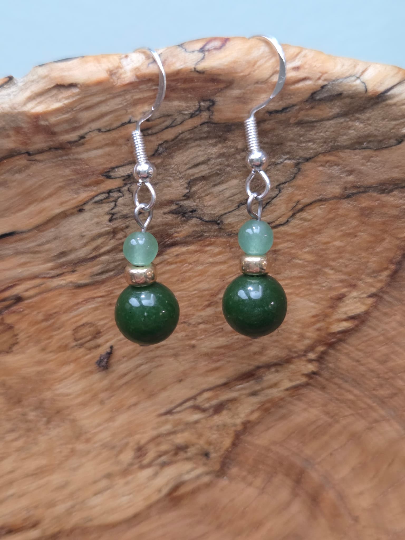Pair of dangle earrings featuring a deep green jade round bead below a lighter sage green aventurine bead, joined by a gold-tone accent bead, on silver-tone fish hook ear wires, resting on a piece of natural wood