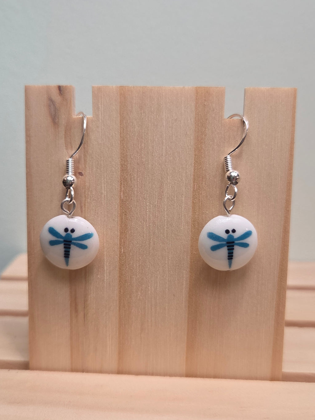 Pair of round white glass bead drop earrings hand-painted with a blue dragonfly design, shown on a wooden ruler stand measuring approximately 1 inch