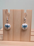 Pair of round white glass bead drop earrings hand-painted with a blue dragonfly design, shown on a wooden ruler stand measuring approximately 1 inch