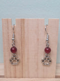 Paw print and mystery stone drop earrings displayed on a wooden earring stand with ruler showing approximately 1.5 inch length