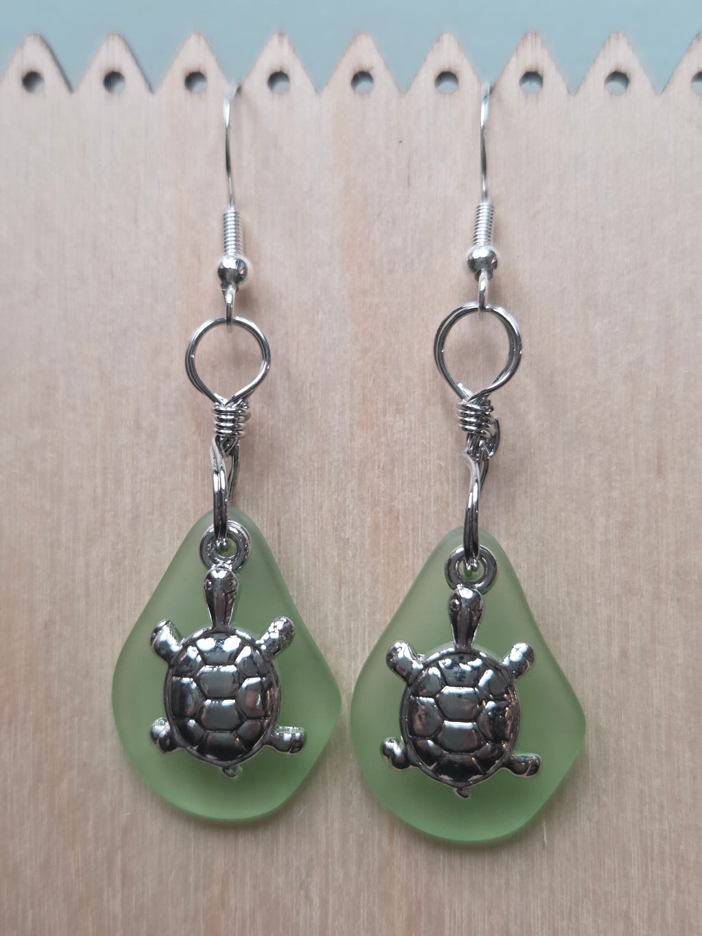 Sage green frosted glass dangle earrings with a silver-tone turtle charm, shown on a white mannequin ear