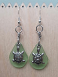 Sage green frosted glass dangle earrings with a silver-tone turtle charm, shown on a white mannequin ear