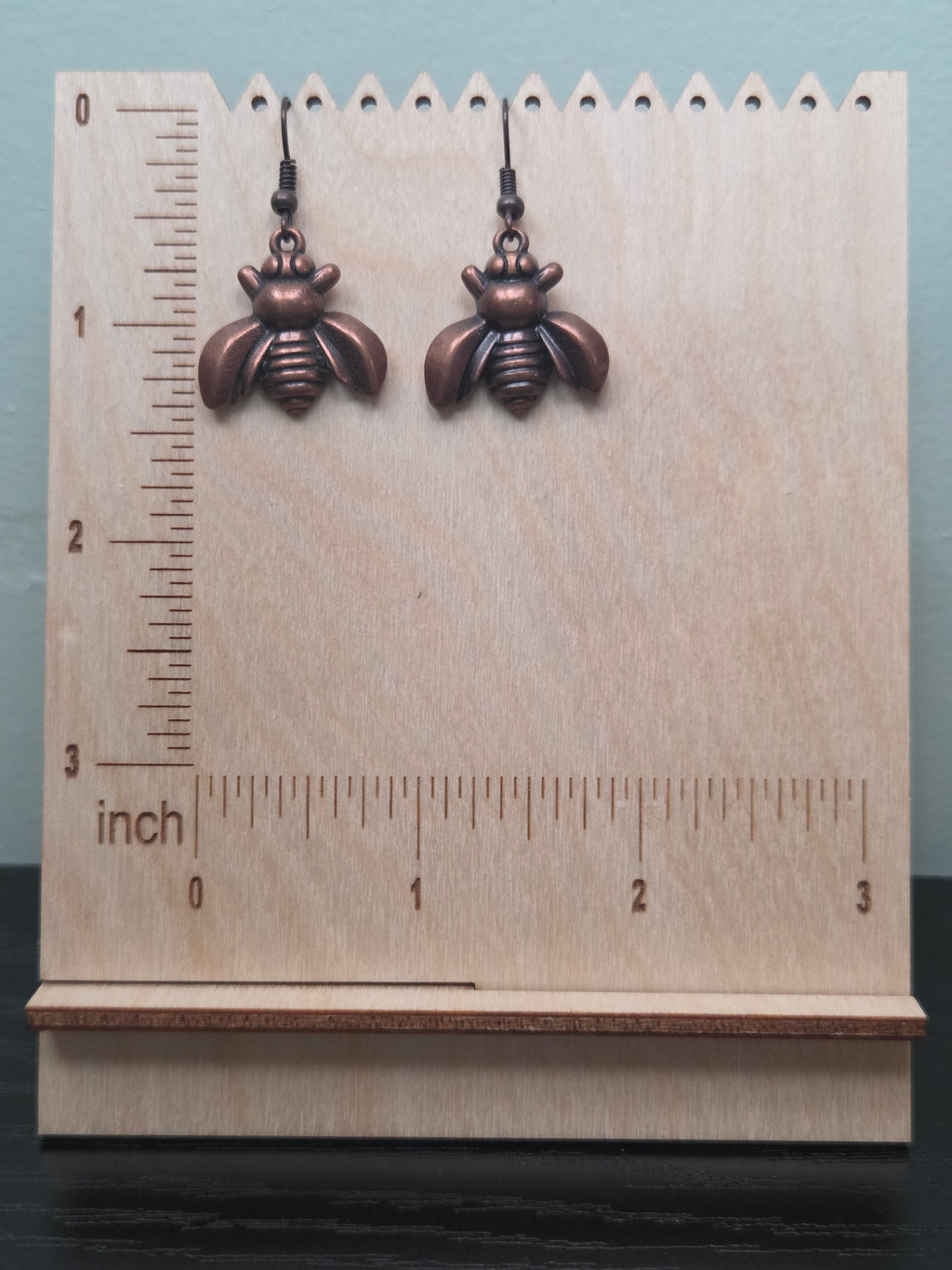 Antiqued copper-tone bee charm dangle earrings against ruler showing approximately 1 inch length