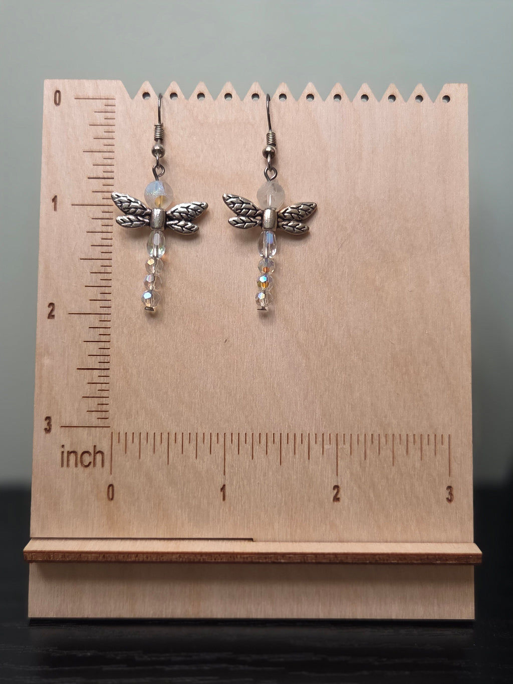Close-up of crystal dragonfly earrings with detailed silver-tone wings and faceted iridescent glass beads shown against a wood block backdrop