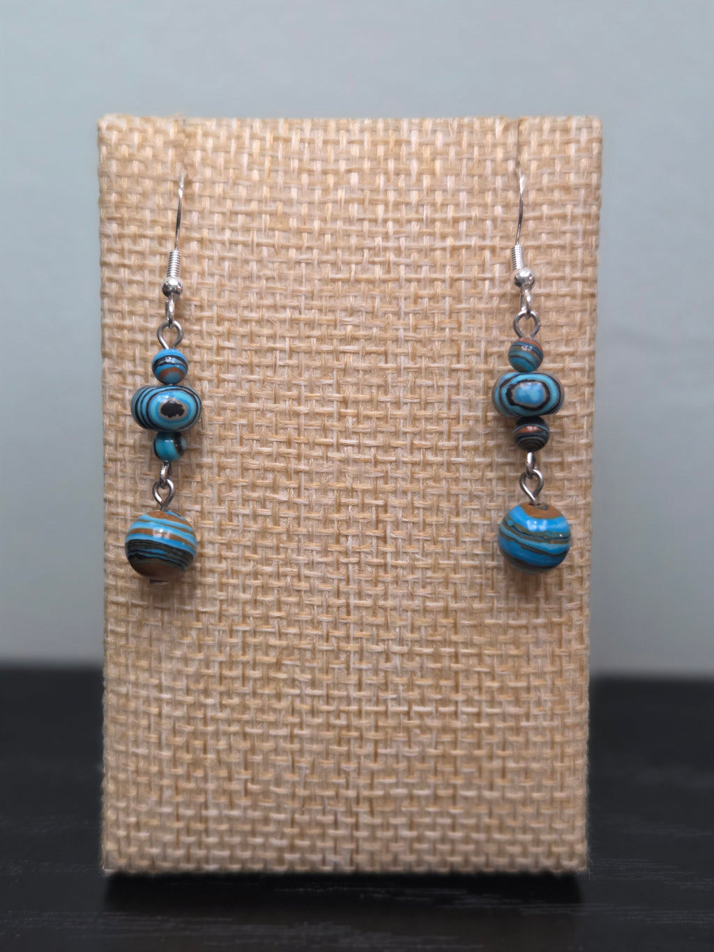 Pair of mystery stone graduated drop earrings with swirled turquoise, black, and tan beads, shown against a woven burlap display card