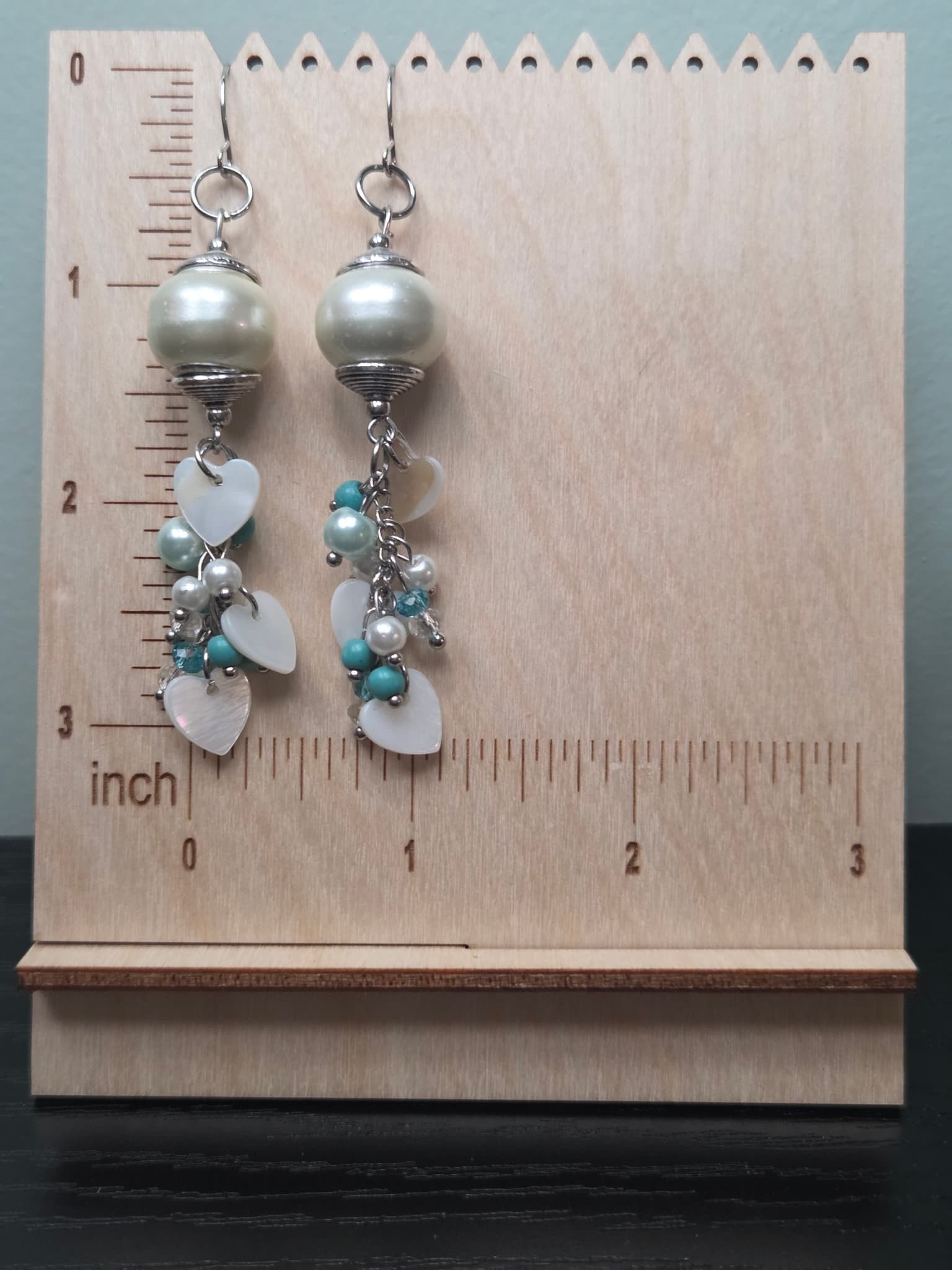 Long dangle drop earrings featuring a large cream pearl bead top with silver wire wrapping, cascading cluster of white mother of pearl heart charms and turquoise and pearl beads, displayed on a wooden earring stand with inch ruler showing approximately 3 inches in length.