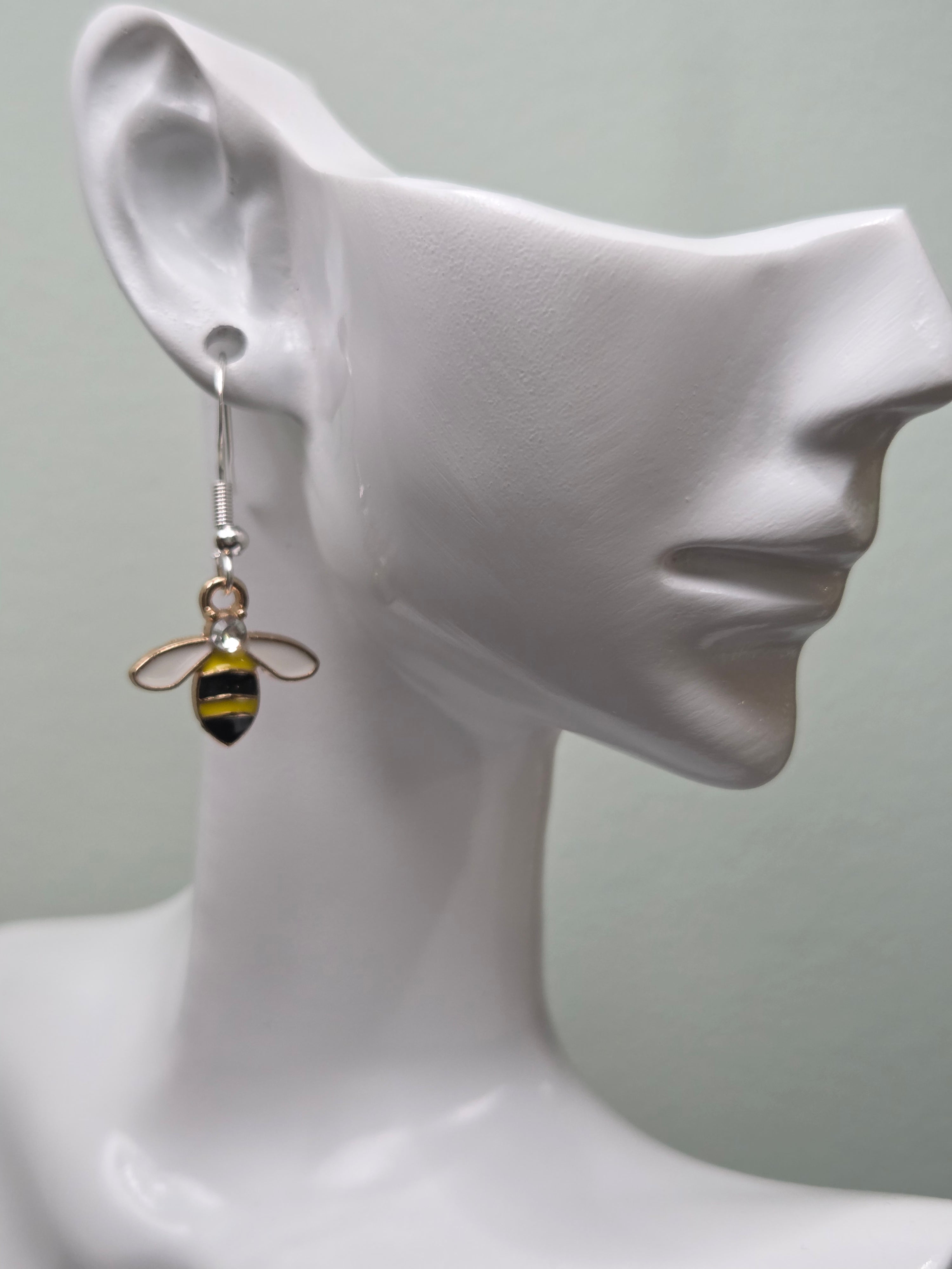 Close-up of a bumble bee dangle earring with yellow and black enamel stripes, clear rhinestone accent, and silver-tone fishhook wire, shown on a white mannequin ear