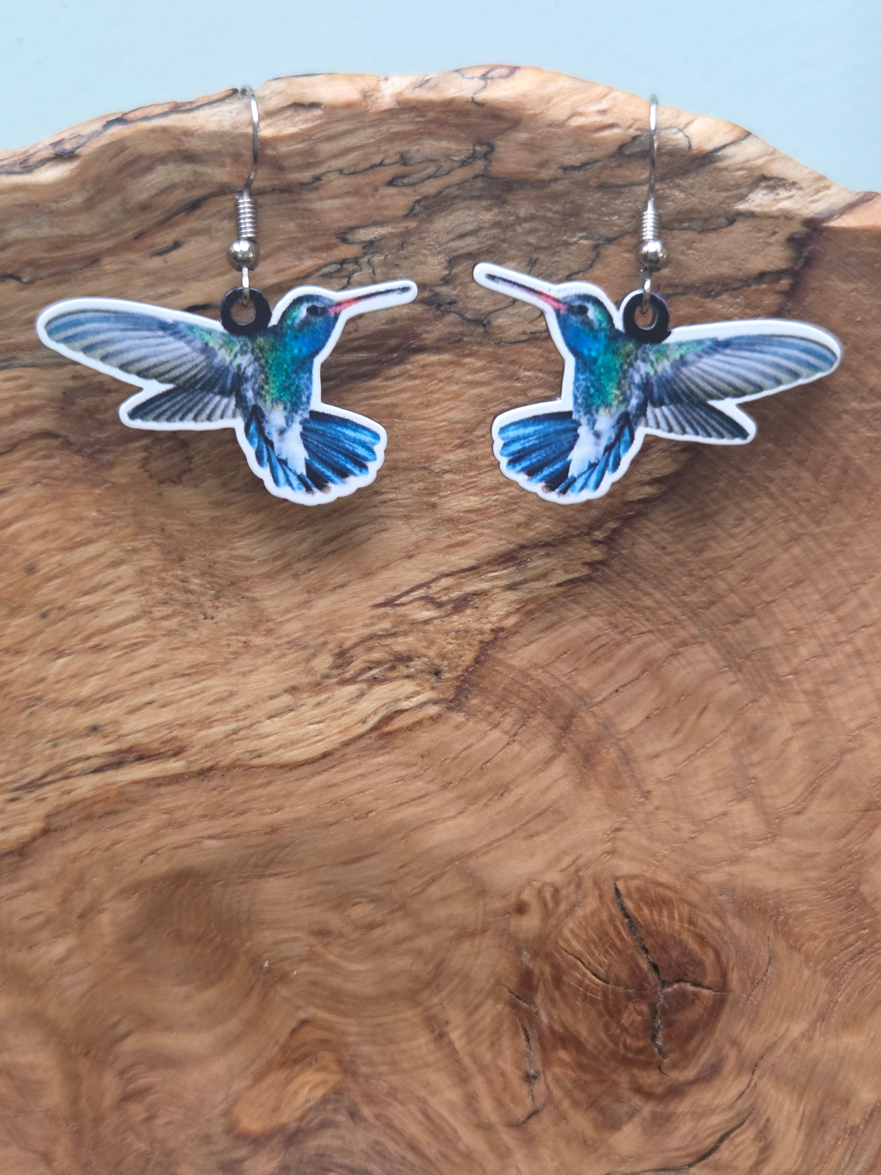 Realistic printed acrylic hummingbird dangle earrings in blue, green, and white, shown on wood display