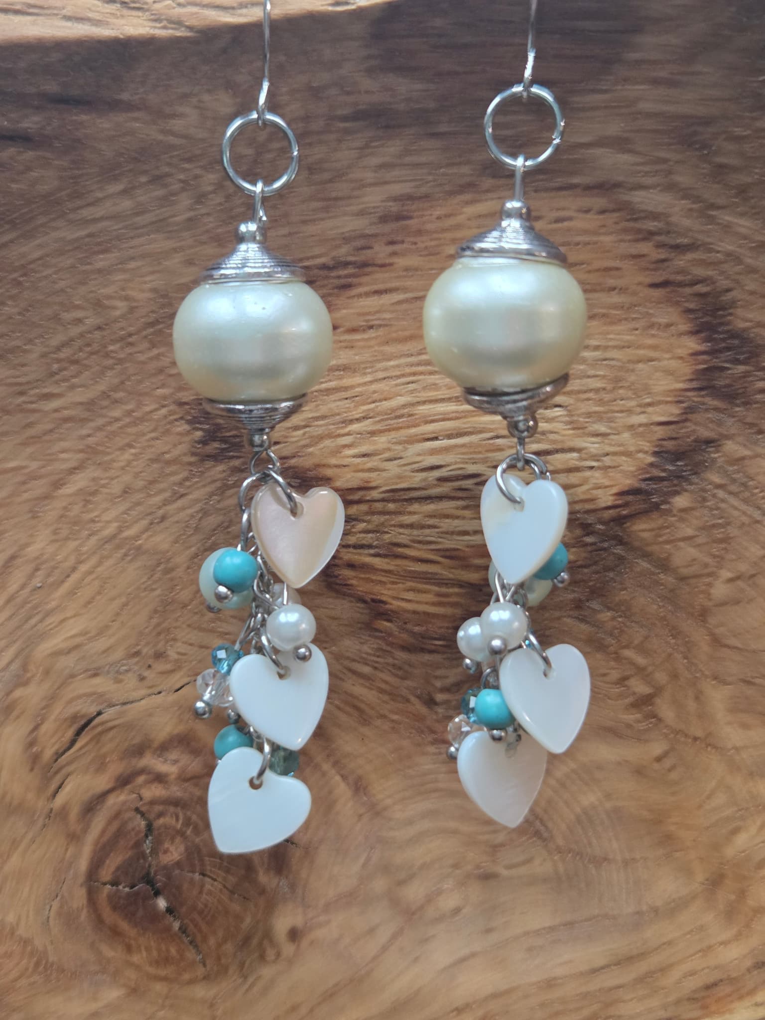 Handmade dangle earrings with a large cream faux pearl bead, silver wire-wrapped cap, and a cascading cluster of white mother of pearl heart charms mixed with turquoise and pearl beads, displayed on a rustic wood grain surface.