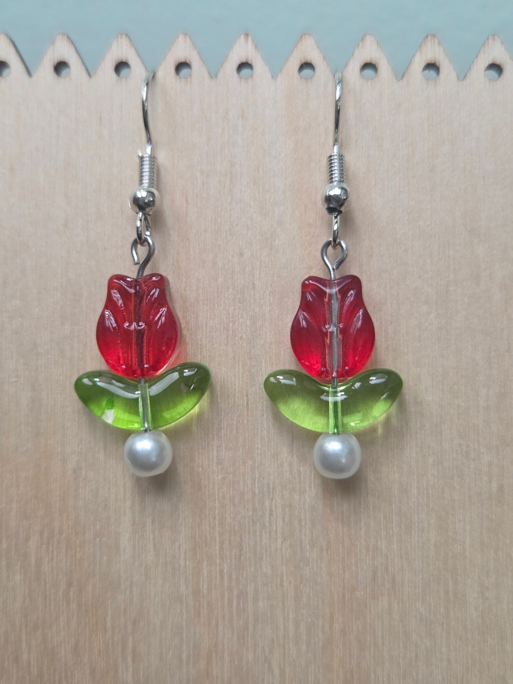 Glass tulip drop earrings with red tulip flower bead, green leaf, and glass pearl accent, displayed on a wooden earring stand with ruler showing approximately 1.75 inch length