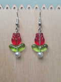 Glass tulip drop earrings with red tulip flower bead, green leaf, and glass pearl accent, displayed on a wooden earring stand with ruler showing approximately 1.75 inch length