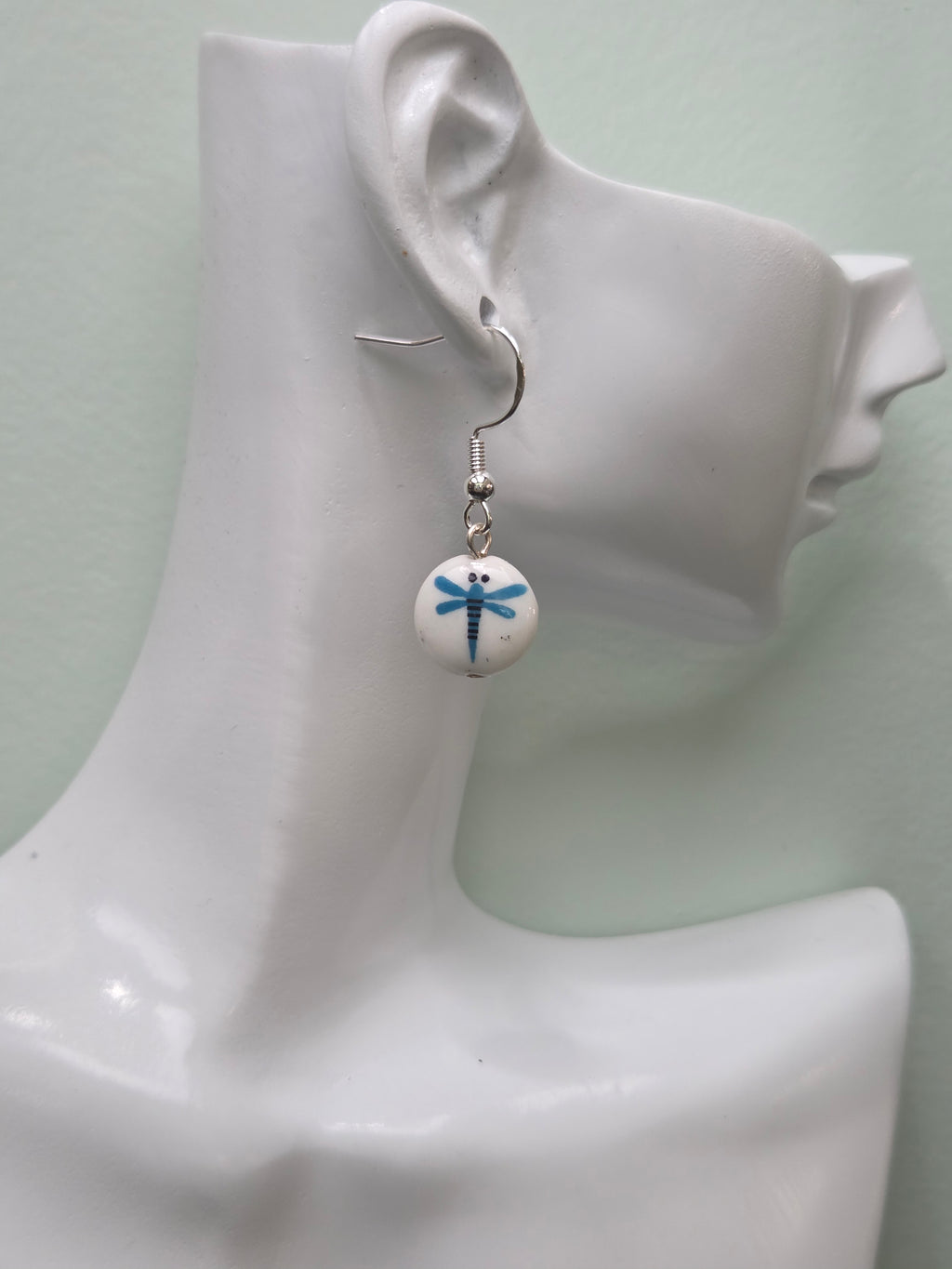 White round glass bead earring with blue painted dragonfly motif, shown on white mannequin ear with silver-tone hook, natural lighting