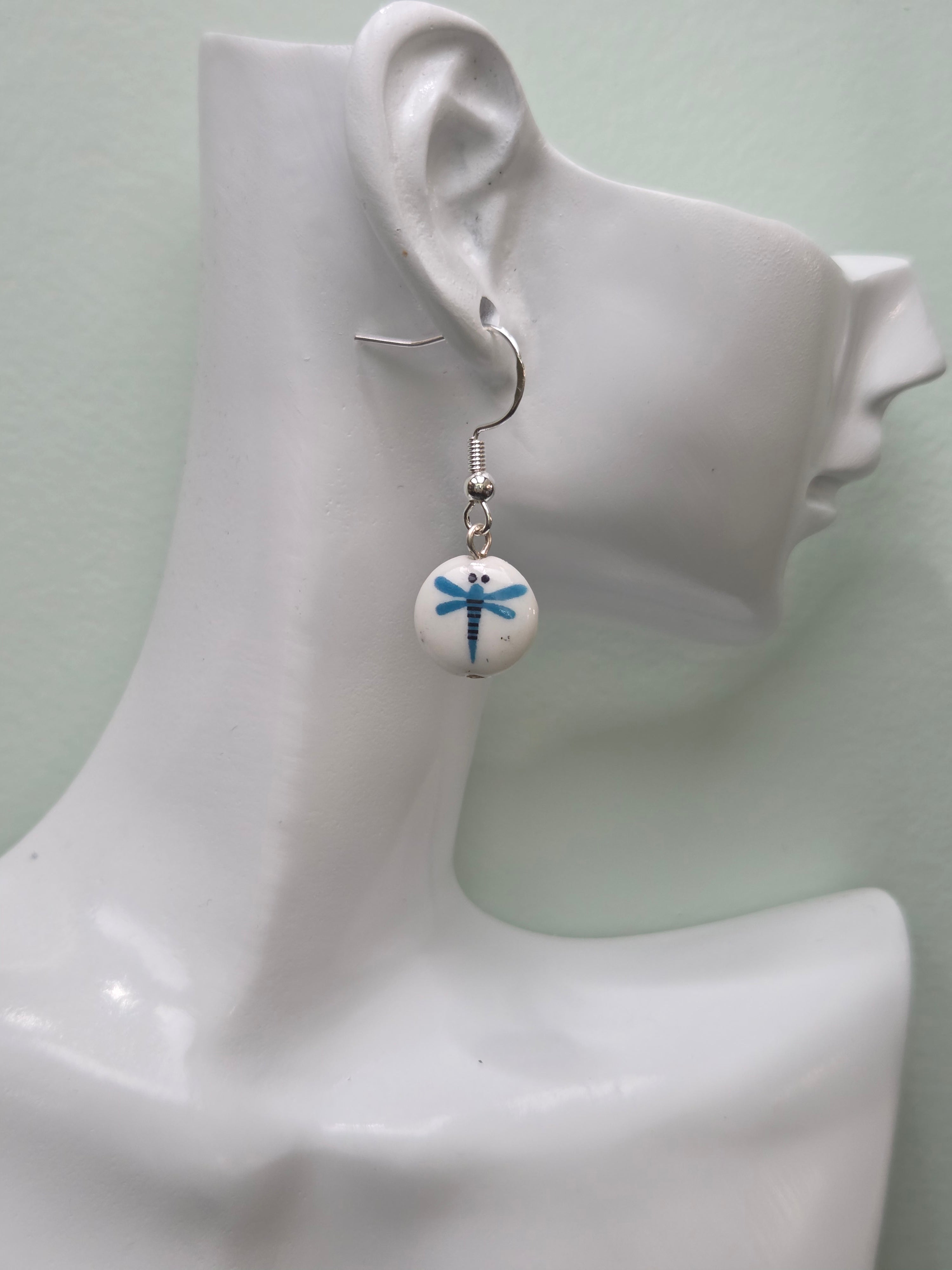 White round glass bead earring with blue painted dragonfly motif, shown on white mannequin ear with silver-tone hook, natural lighting