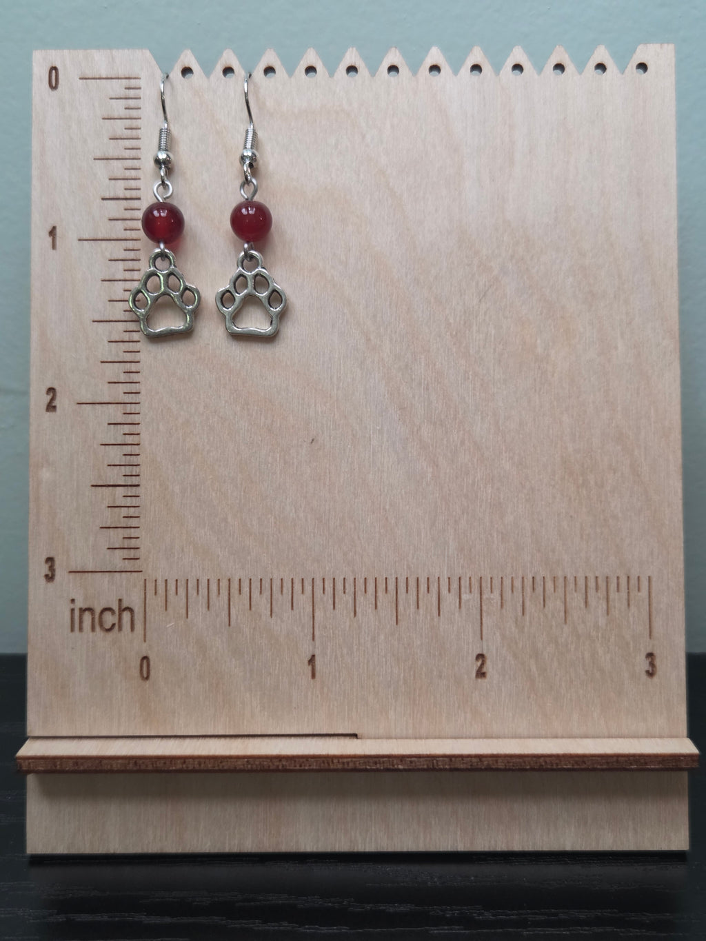 Silver-tone paw print charm earrings with deep red mystery stone bead, shown on natural wood background