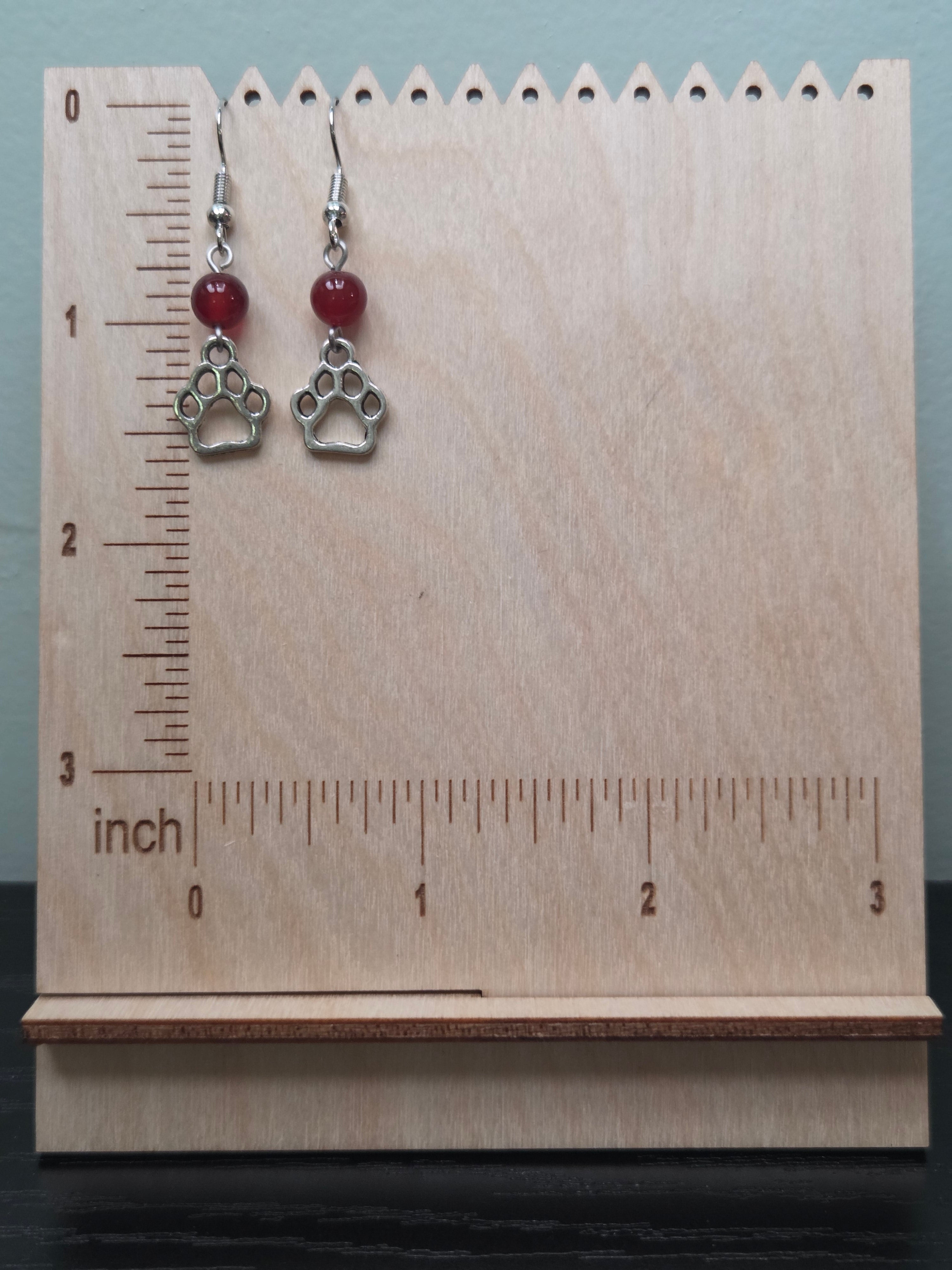 Silver-tone paw print charm earrings with deep red mystery stone bead, shown on natural wood background